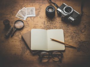A sepia-toned scene photographed from above shows a magnifying glass, some photographs, a camera and lens, and an open notebook with a pencil laid across it. A pair of glasses lie in front of the notebook.