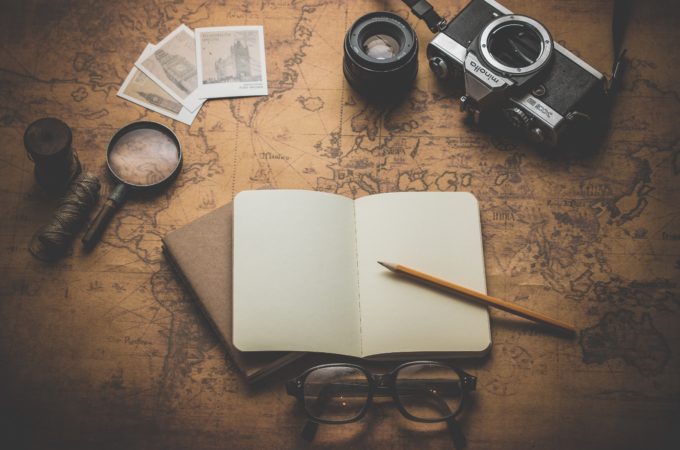 A sepia-toned scene photographed from above shows a magnifying glass, some photographs, a camera and lens, and an open notebook with a pencil laid across it. A pair of glasses lie in front of the notebook.