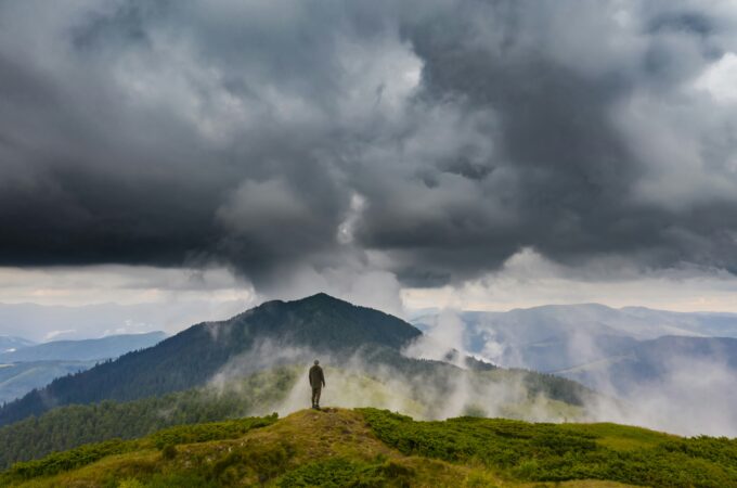 A storm approaches. The vista of mountains and a man standing upon a mountain that the storm has not yet reached.
