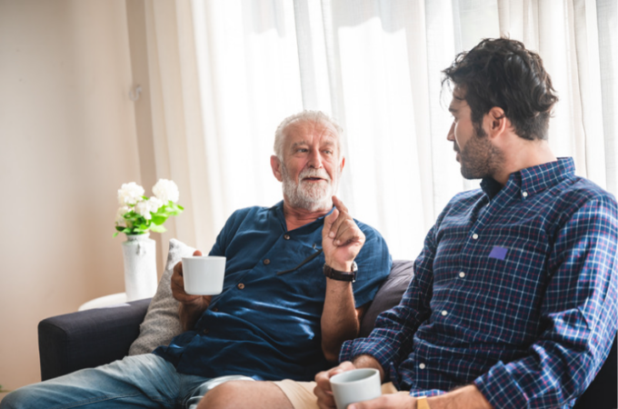 Father speaks to son. They are sitting on a couch in a living room. Father gesticulates to son.