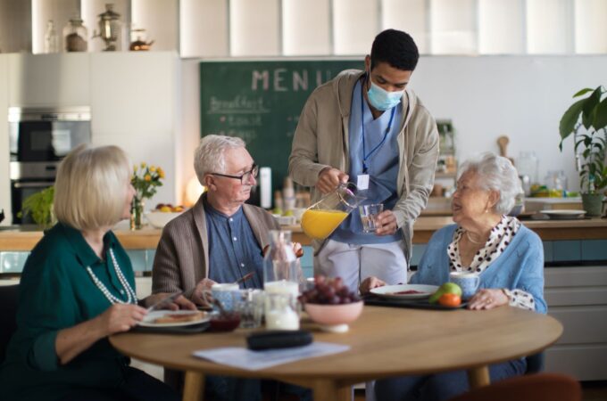 Seniors have breakfast together. They are served by a man in health care scrubs.